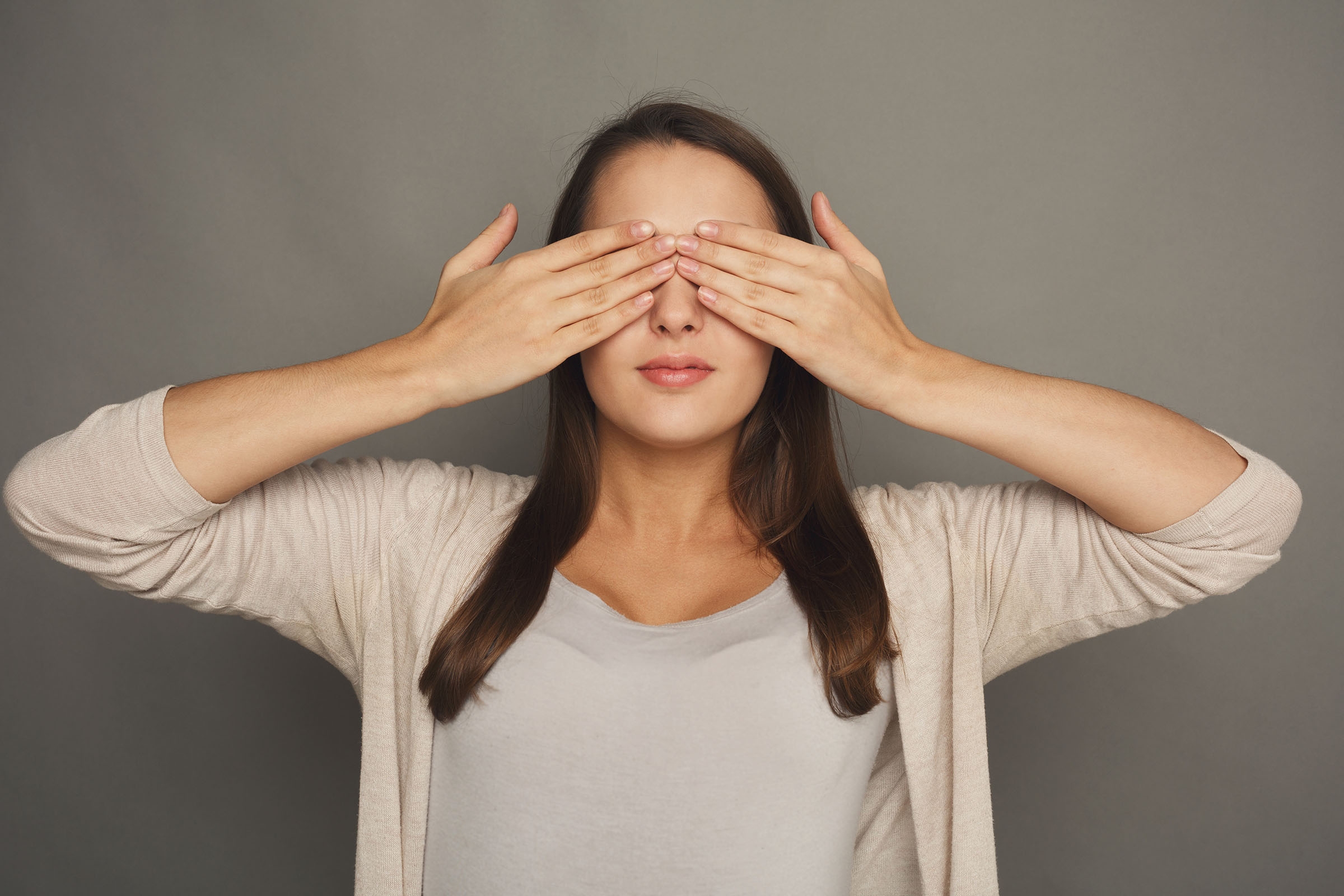 See no evil concept. Portrait of young scared woman covering eyes with hands, gray studio background.